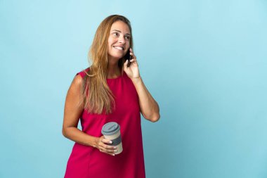 Mavi arka planda izole edilmiş orta yaşlı Brezilyalı kadın elinde kahve ve cep telefonu var.