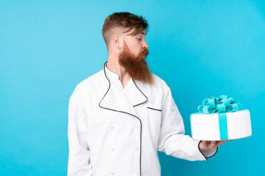 Kızıl saçlı, uzun sakallı bir pasta şefi izole edilmiş mavi arka planda mutlu bir ifadeyle büyük bir pasta tutuyor.