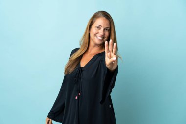 Mavi arka planda izole edilmiş orta yaşlı Brezilyalı kadın mutlu ve parmaklarıyla üçe kadar sayıyor.