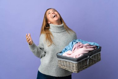 Mor arka planda izole edilmiş çamaşır sepetini tutan orta yaşlı kadın gülüyor.