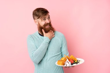 Kızıl saçlı, uzun sakallı, pembe arka planda waffle tutan bir adam.