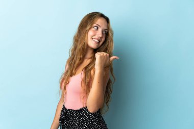 Mavi arka planda izole edilmiş genç sarışın kadın bir ürünü sunmak için kenarı işaret ediyor.