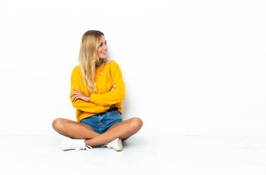 Genç sarışın Uruguaylı kadın, beyaz arka planda izole bir şekilde, kolları bağlı ve mutlu bir şekilde yerde oturuyor.