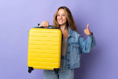 Genç sarışın kadın, mor arka planda izole edilmiş tatilde seyahat valiziyle ve başparmağıyla