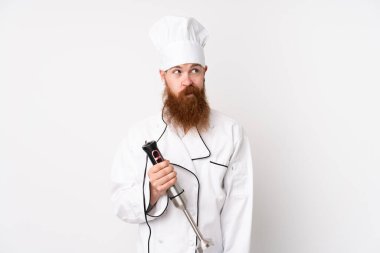 Kızıl saçlı adam el blenderi kullanıyor. İzole edilmiş beyaz arka plan üzerinde duruyor ve yan tarafa bakıyor.