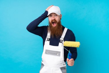 Uzun sakallı ressam mavi arka planda şaşırmış ve parmağıyla kenarı işaret ediyor.