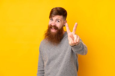 Uzun sakallı kızıl saçlı adam izole edilmiş sarı arka planda gülümsüyor ve zafer işareti gösteriyor.
