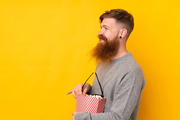 Redhead man with long beard over isolated yellow background holding a big bucket of popcorns