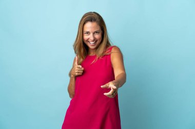 Mavi arka planda izole edilmiş orta yaşlı Brezilyalı kadın ön tarafı işaret ediyor ve gülümsüyor.