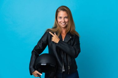 Mavi arka planda izole edilmiş motosiklet kaskı tutan orta yaşlı bir kadın bir ürünü sunmak için kenarı işaret ediyor.