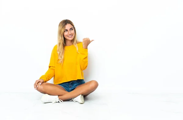 Jonge Blonde Uruguayaanse Vrouw Zit Vloer Geïsoleerd Witte Achtergrond Wijzend — Stockfoto