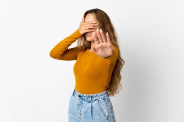 Mavi arka planda izole edilmiş genç sarışın kadın dur hareketi yapıyor ve yüzünü kapatıyor.