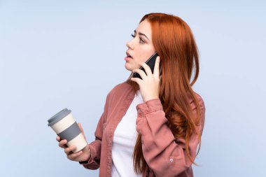 İzole edilmiş mavi arka planı olan kızıl saçlı genç kız elinde kahve ve bir cep telefonu tutuyor.