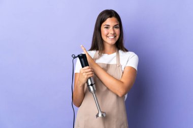 Mor arka planda izole edilmiş el blenderi kullanan Brezilyalı kadın bir ürünü sunmak için yan tarafı işaret ediyor