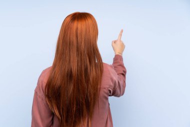 İzole edilmiş mavi arka planı olan kızıl saçlı genç kız işaret parmağıyla işaret ediyor.