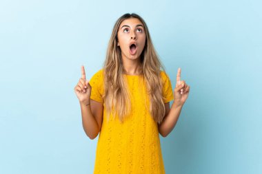 Tecrit edilmiş mavi arka planı olan genç İspanyol kadın şaşırmış ve işaret ediyor.