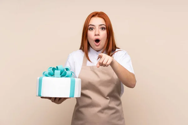 Redhead teenager girl with a big cake over isolated background surprised and pointing front