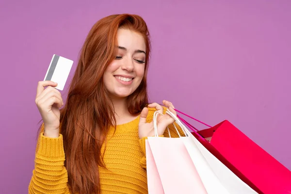 Redheadティーン女の子上の隔離された紫背景保持ショッピングバッグとクレジットカード — ストック写真