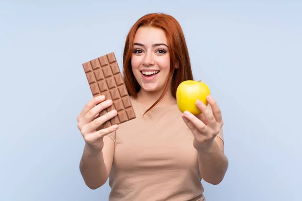 赤毛ティーン女の子上の隔離された青の背景取るAチョコレートタブレットで一方でと一つのアップルでもう一方 — ストック写真