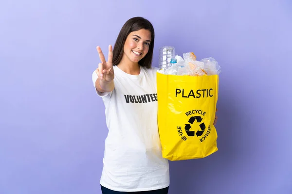 若いブラジル人女性は 紫色の背景笑顔で孤立したリサイクルし 勝利の兆候を示すためにビニールボトルでいっぱいの袋を保持 — ストック写真