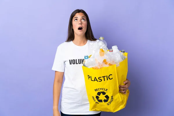 紫色の背景に隔離されたリサイクルのためにビニールボトルでいっぱいの袋を持っている若いブラジル人女性が見上げと驚きの表情で — ストック写真