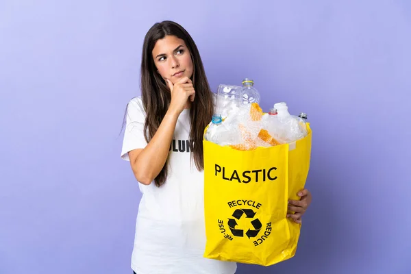 若いブラジル人女性は袋を持っていますプラスチックボトルでいっぱい紫色の背景に隔離されたリサイクル疑問を持っています — ストック写真