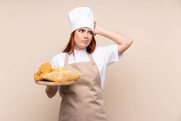 红头发少女穿着厨师制服 女面包师拿着桌子 几个面包 满脸疑惑 表情混乱 — 图库照片