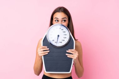Tartı makinesi olan güzel bir genç kız izole edilmiş pembe arka planda tartı makinesiyle arkasına saklanıyor.