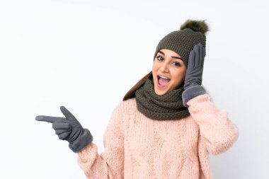 Kışlık şapkalı genç kız izole edilmiş beyaz arka planda şaşırmış ve parmağıyla kenarı işaret ediyor.