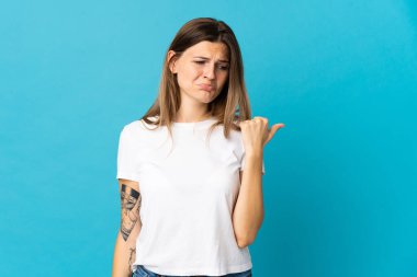 Genç Slovak kadın mavi arka planda izole edilmiş mutsuz ve kenarı işaret ediyor.