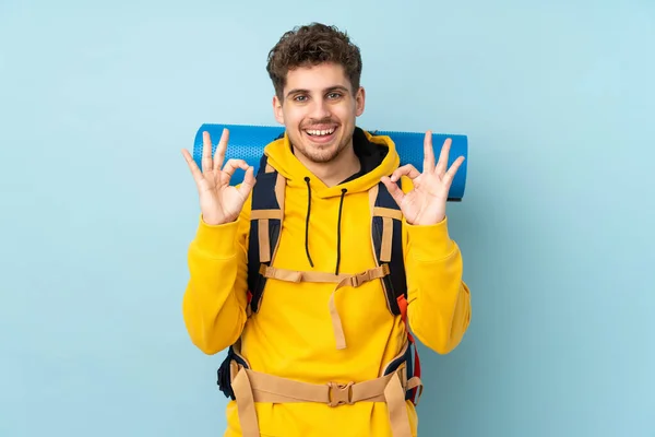 若いです登山家男とともに大きなバックパックで隔離された青の背景を示します指でOkサイン — ストック写真