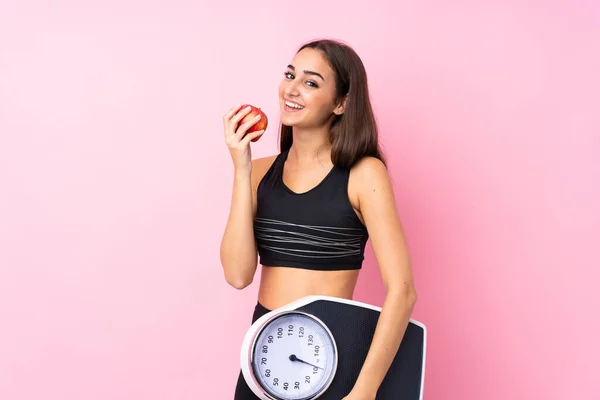 Pretty young girl with weighing machine over isolated pink background with weighing machine and with an apple