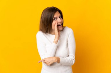 Sarı arka planda izole edilmiş genç Slovak kadın bir ürün sunmak ve bir şeyler fısıldamak için kenarı işaret ediyor.