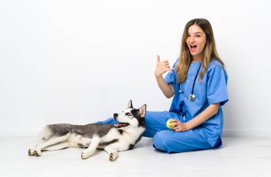 Sibiryalı Husky köpeği olan veteriner doktor yerde oturmuş, telefon işareti yapıyor.
