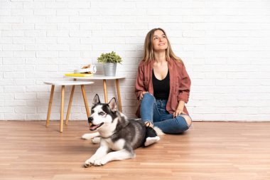 Köpeğiyle yerde oturan genç bir kız. Kapalı alanda yukarı bakıp gülümsüyor.