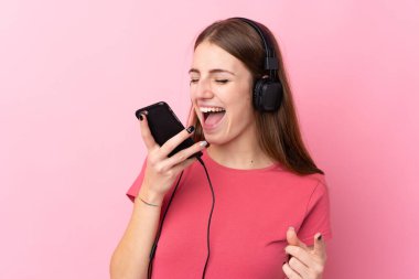 Soyutlanmış pembe arka planı olan genç bir kadın cep telefonuyla müzik dinliyor ve şarkı söylüyor.