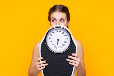 Arkasında tartı makinesi olan, izole edilmiş sarı arka planı olan genç bir kadın.