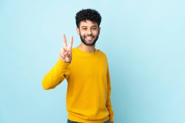 Mavi arka planda izole edilmiş genç Faslı adam gülümsüyor ve zafer işareti gösteriyor.