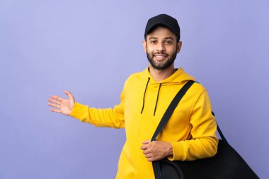 Spor çantalı genç Faslı adam mor arka planda izole edilmiş. Davet etmek için ellerini yana uzatıyor.
