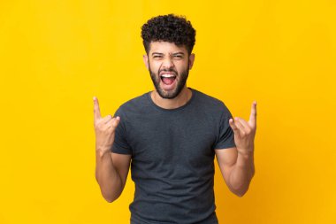 Sarı arka planda izole edilmiş genç Faslı adam boynuz hareketi yapıyor.