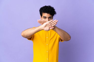 Genç Venezuelalı bir adam mor arka planda izole edilmiş bir şekilde bir eylemi durdurmak için el hareketi yapıyor.