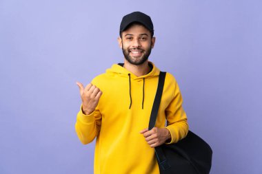 Mor arka planda izole edilmiş spor çantalı genç Faslı bir adam bir ürünü sunmak için kenarı işaret ediyor.