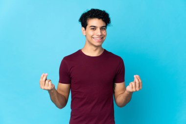 Genç Venezuelalı adam mavi arka planda izole edilmiş para hareketi yapıyor.