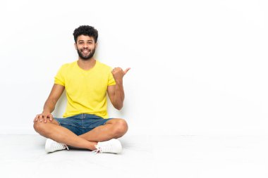 Genç Faslı yakışıklı bir adam izole bir zeminde oturuyor ve bir ürünü sunmak için yan tarafı işaret ediyor.