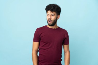 Faslı genç bir adam mavi arka planda izole edilmiş. Yan tarafa bakarken sürpriz jest yapıyor.