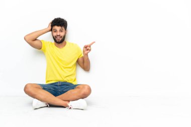 Genç Faslı yakışıklı adam izole bir arka planda yerde oturuyor. Şaşırmış ve parmağıyla işaret ediyor.