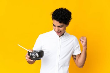 Genç Venezuelalı bir adam sarı arka planda izole edilmiş bir şekilde zaferini kutlarken elinde yemek çubuklarıyla bir kase erişte tutuyordu.