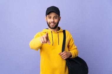 Spor çantalı genç Faslı adam, mor arka planda izole edilmiş, şaşırmış ve ön tarafı işaret ediyor.