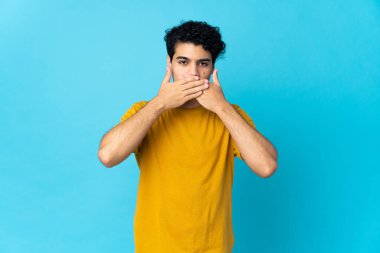 Genç Venezuelalı adam mavi arka planda izole edilmiş, ağzı elleriyle kapatıyor.