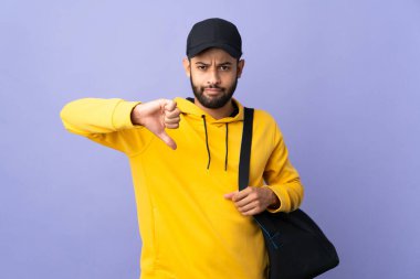 Spor çantalı genç Faslı adam, mor arka planda izole edilmiş, olumsuz ifadeli baş parmağını gösteriyor.